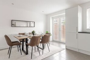 Un espace repas moderne avec une table dressée pour quatre personnes, entourée de chaises, et une cuisine dotée de meubles blancs et d’une fenêtre.