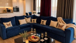 A modern living room with a blue sectional sofa, a coffee table with fruit and glasses, and a kitchen in the background. - Southampton 2-Bed Apartment, Sleeps 4 with Balcony (Southampton)