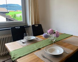 Una mesa de comedor dispuesta con platos, cubiertos y un florero, con vistas a una casa y vegetación.