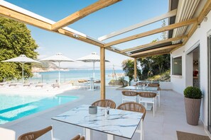 An outdoor pool area with a glass table and chairs, surrounded by umbrellas and lounge chairs, overlooking a body of water. - Alboro Seaside Suites (Parga)