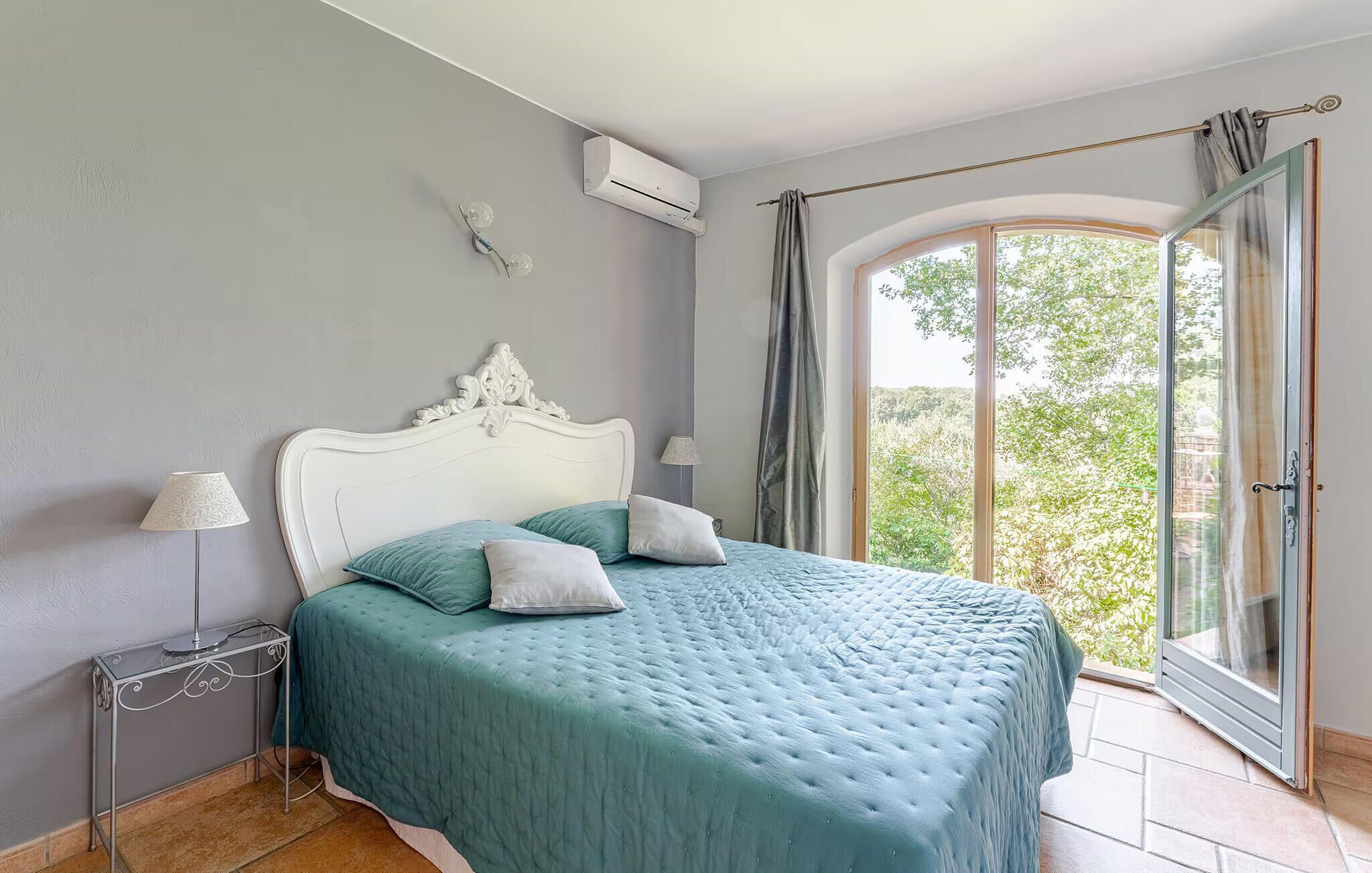 Una camera da letto con un letto, un comodino, un condizionatore, una finestra con tende e vista sul verde.