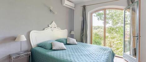 Un dormitorio con cama, mesita de noche, equipo de aire acondicionado, ventana con cortinas y vista a vegetación exterior.