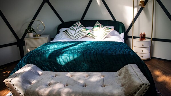 A bed with a teal quilt, a tufted bench, a bedside table with a lamp, and a wall with a geometric pattern.