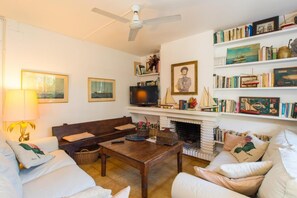 A living room with a sofa, a coffee table, a television, and a fireplace. There are bookshelves with books and framed pictures on the wall. - O-1 Beautiful ground floor close to the beach and with pool (Calella de Palafrugell)