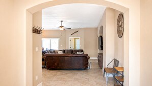 A spacious living room with a brown leather couch, a ceiling fan, and a large clock on the wall.