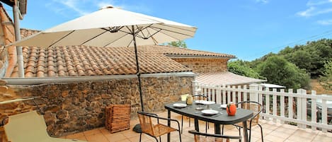 Een terras buiten met een tafel, stoelen, een witte parasol en een stenen muur.