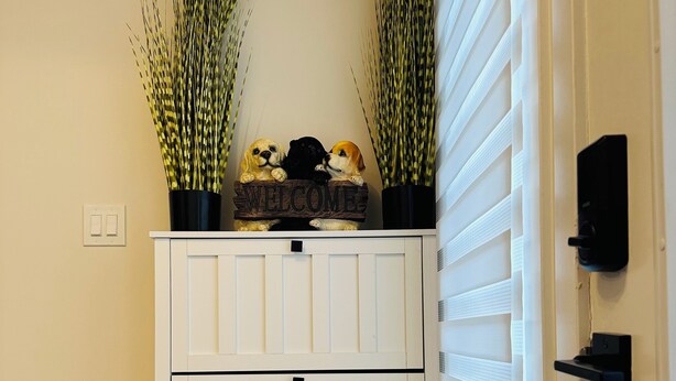 Un mueble blanco con un cartel que dice 'WELCOME' y adornos de perros, flanqueado por plantas altas, sobre un muro beige con una ventana a la derecha.