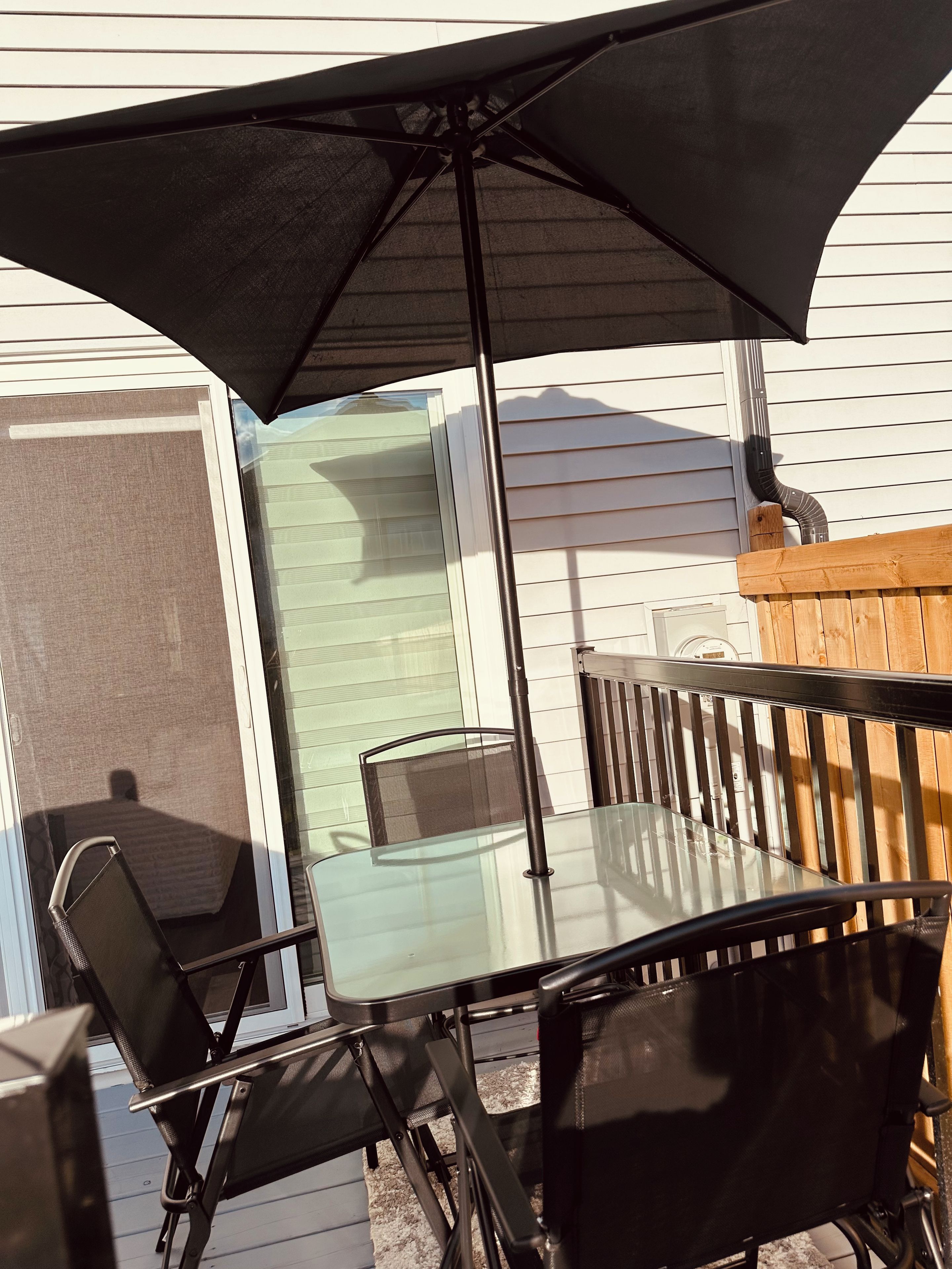An outdoor patio with a glass table and chairs, a black umbrella, and a wooden fence.