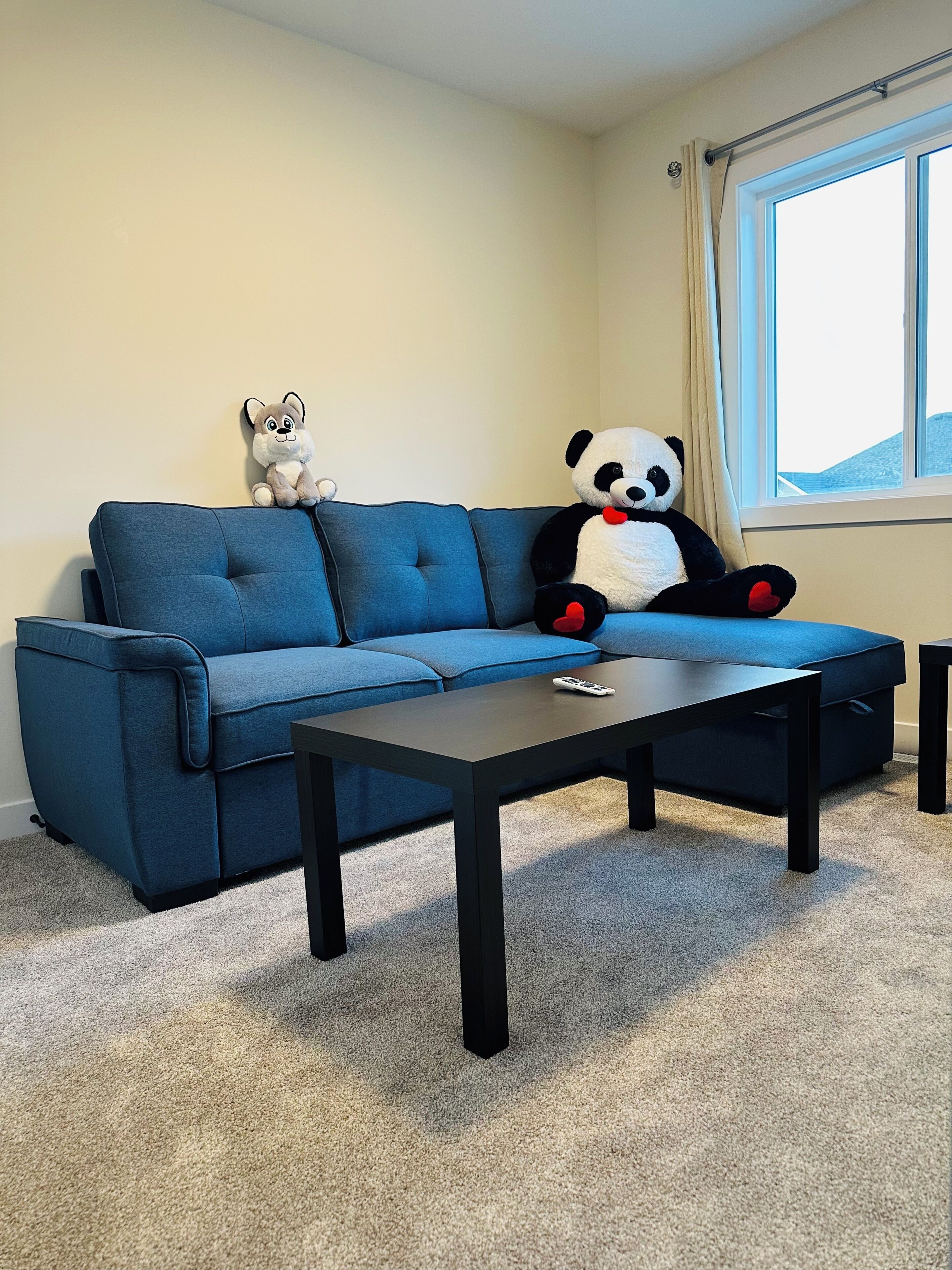 A living room with a blue sofa, a small dog, a large panda plush toy, and a remote control on the coffee table.
