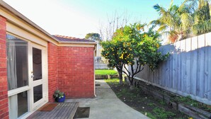 A backyard with a red brick house, a wooden deck, a wooden fence, and a tree with yellow fruit. - Large, spacious group stay, quiet area with a private yard, central in Melb S/E (Glen Waverley)