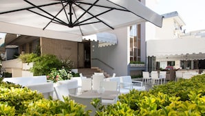 An outdoor seating area with white furniture and a large umbrella, surrounded by greenery. - Hotel Riviera (Jesolo)