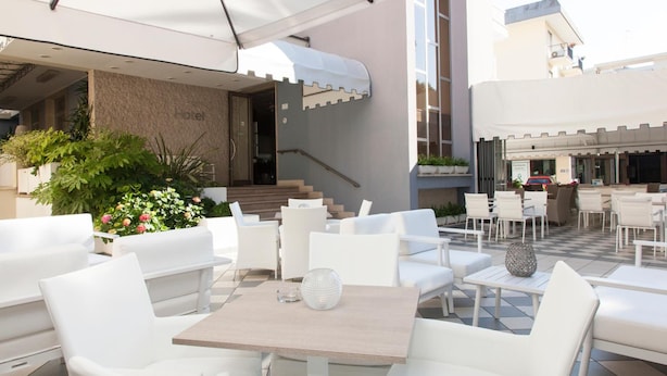 An outdoor seating area with white chairs and tables, a staircase leading to an entrance, and a view of the street. - Hotel Riviera (Jesolo)