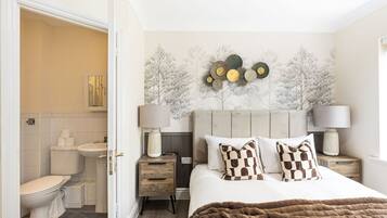 A bedroom with a bed, bedside tables, a nightstand, a lamp, a wall with a tree pattern, and a bathroom visible through an open door.