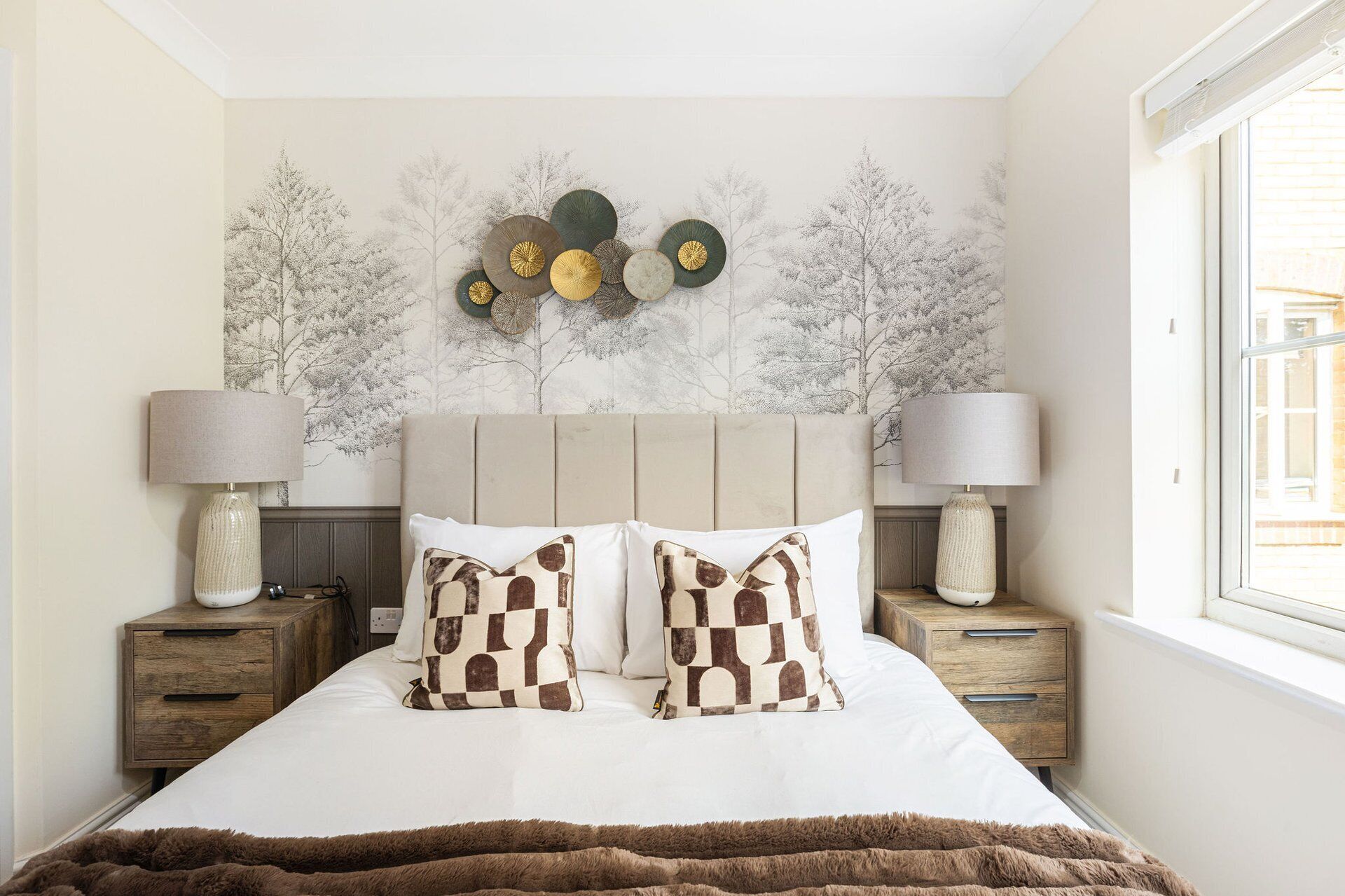 Ein Schlafzimmer mit einem Bett, zwei Nachttischen, zwei Lampen, einem Fenster und einer Wand mit Baum-Motiv.