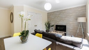 Una sala de estar con sofá, una mesa de comedor con sillas, televisor, planta y lámpara.