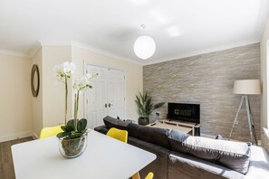 Ein Wohnzimmer mit einer Couch, einem Esstisch mit Stühlen, einem Fernseher, einer Pflanze und einer Lampe.