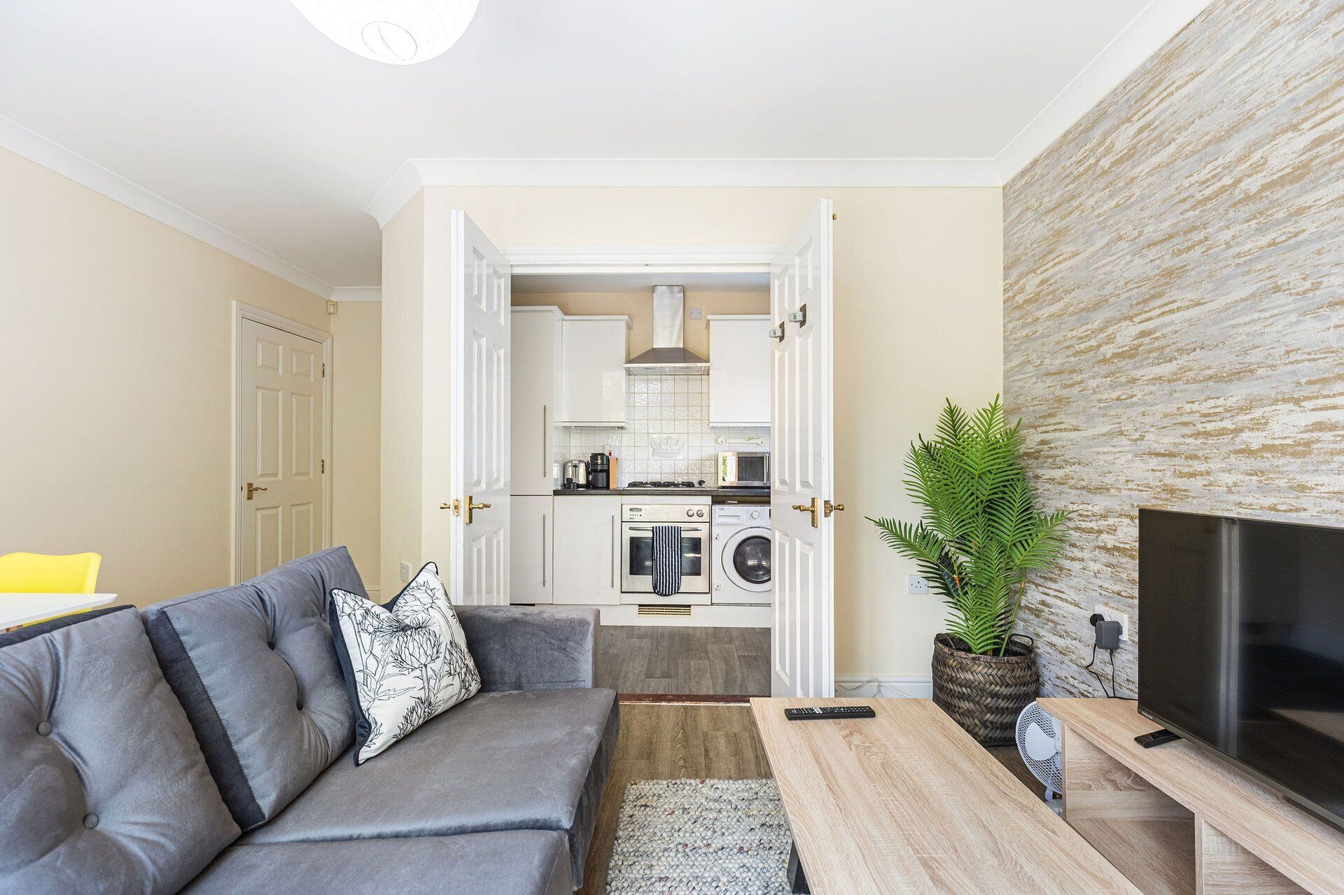 A modern living room with a grey sofa, a flat-screen TV, and a kitchen in the background.
