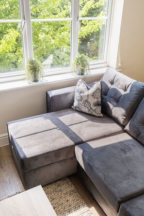 A sectional sofa with a grey fabric, a patterned pillow, and a small potted plant on a windowsill. - GuestReady - Chic Retreat in Harrow (London)