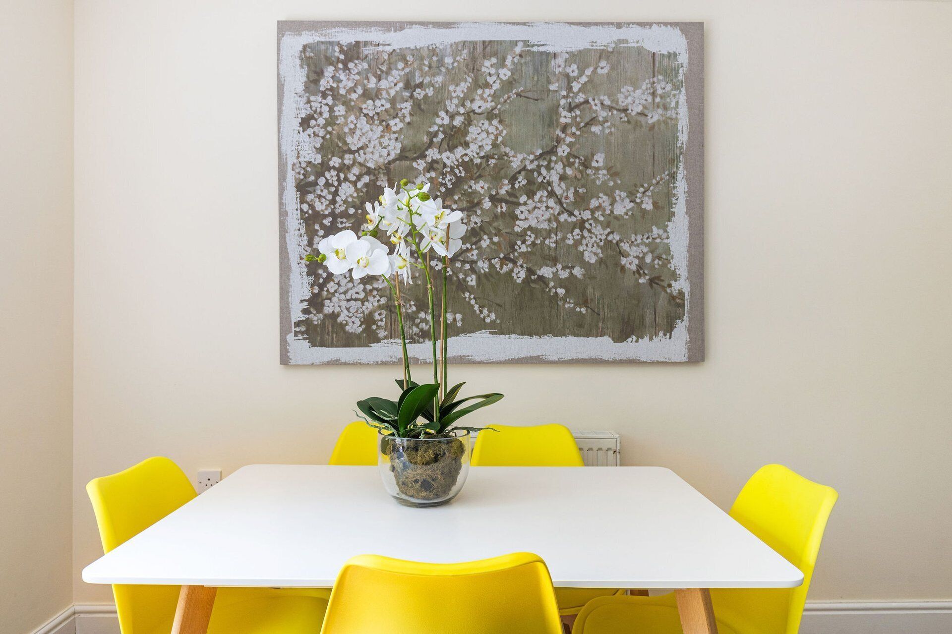 A dining area with a white table and yellow chairs, a potted plant with white flowers, and a large abstract painting on the wall.