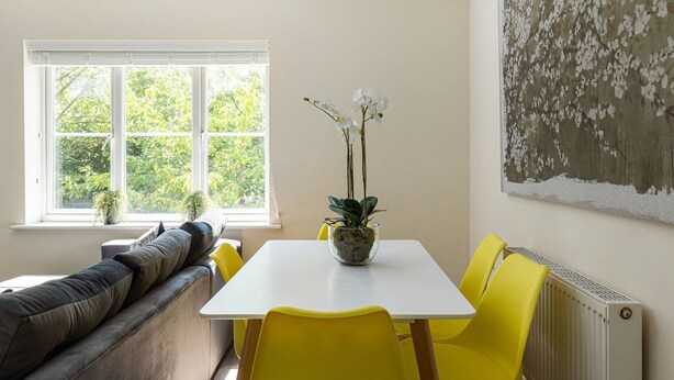 Un comedor con una mesa blanca y sillas amarillas, una ventana con persianas, un sofá y una pared con un cuadro floral.