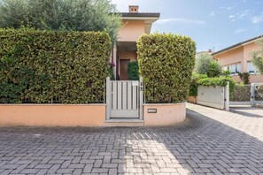 Una entrada con portón y un camino pavimentado que conduce a un edificio de fachada color beige y un pequeño porche.
