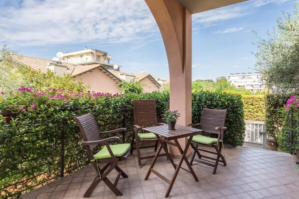 Un patio al aire libre con una mesa y cuatro sillas, rodeado de follaje y edificios al fondo.