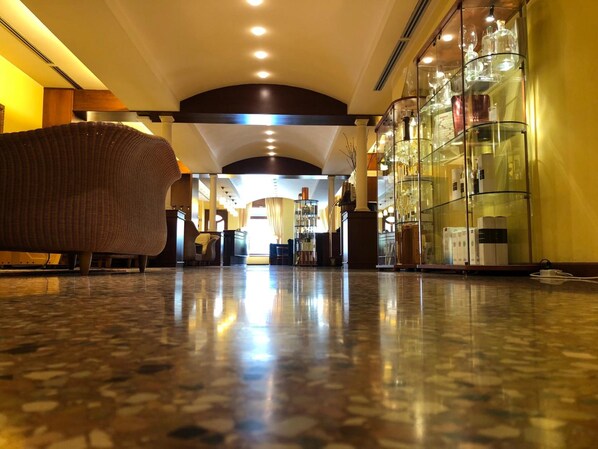 Um saguão de hotel com piso de mármore polido, um sofá de vime e uma vitrine de vidro com diversos objetos.