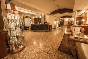 Um hall de hotel com balcão de recepção, uma vitrine de garrafas de vidro, uma área de estar com sofá e poltronas, e um tapete estampado.