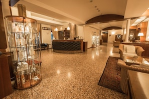 A hotel lobby with a reception desk, a glass display of bottles, a seating area with a sofa and armchairs, and a patterned rug. - La Pieve Hotel (Chiampo)
