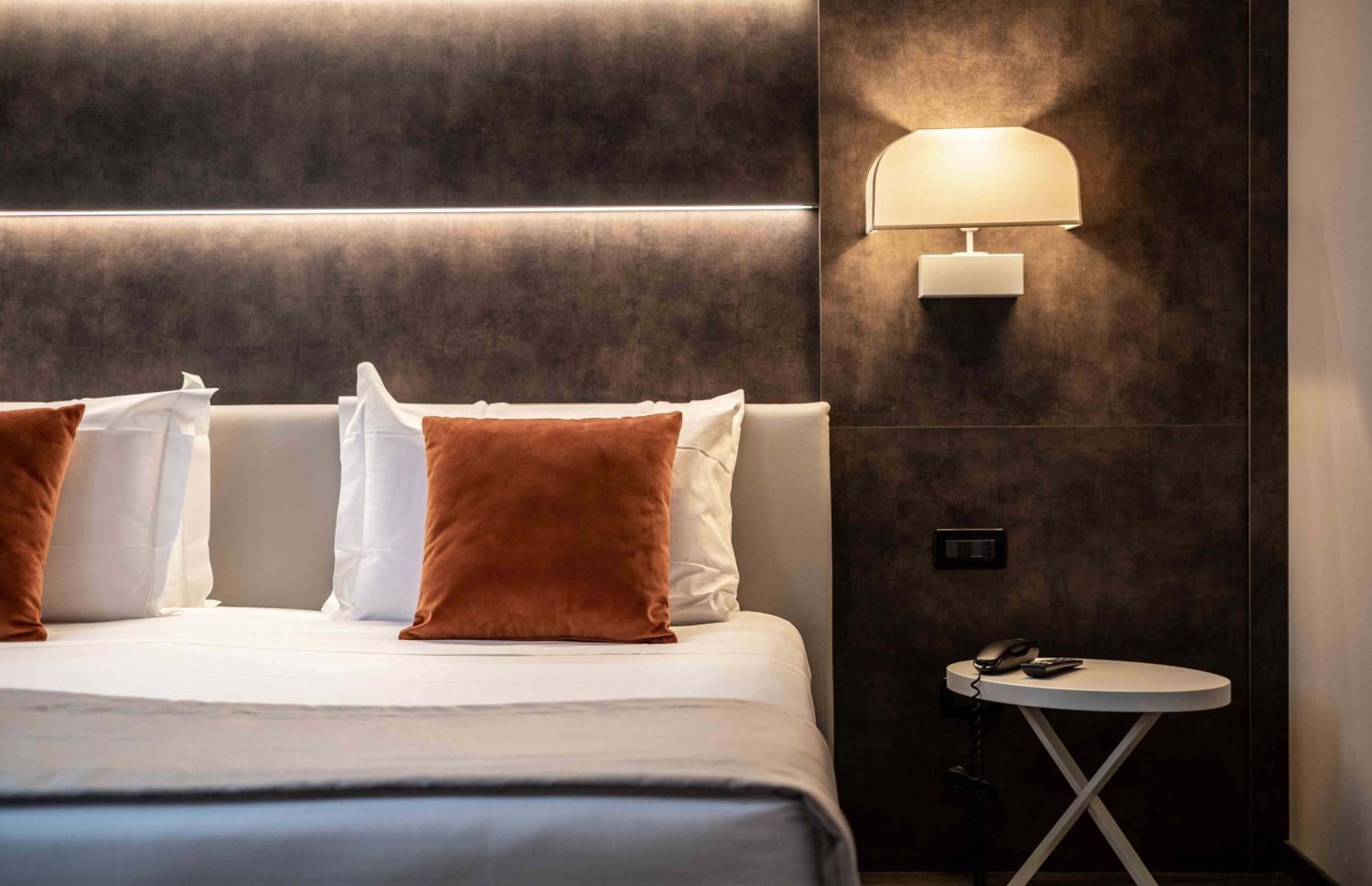 A hotel room with a bed, pillows, a bedside table with a phone, and a wall-mounted lamp.