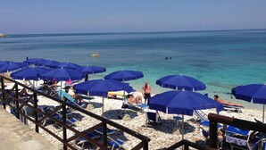 A beach with clear blue water, sun loungers, and blue umbrellas. - Hotel Villa Ombrosa (Portoferraio)