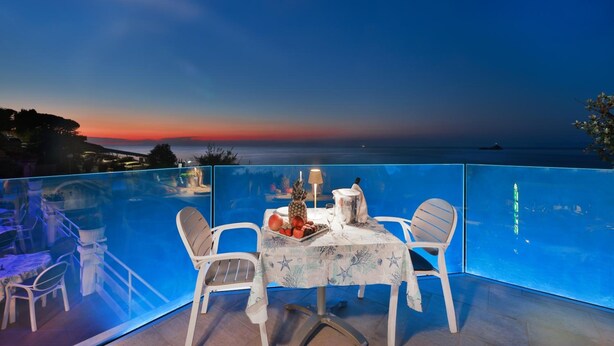 An outdoor dining area with a table set for two, overlooking a scenic view of the sea and a sunset. - Hotel Villa Ombrosa (Portoferraio)