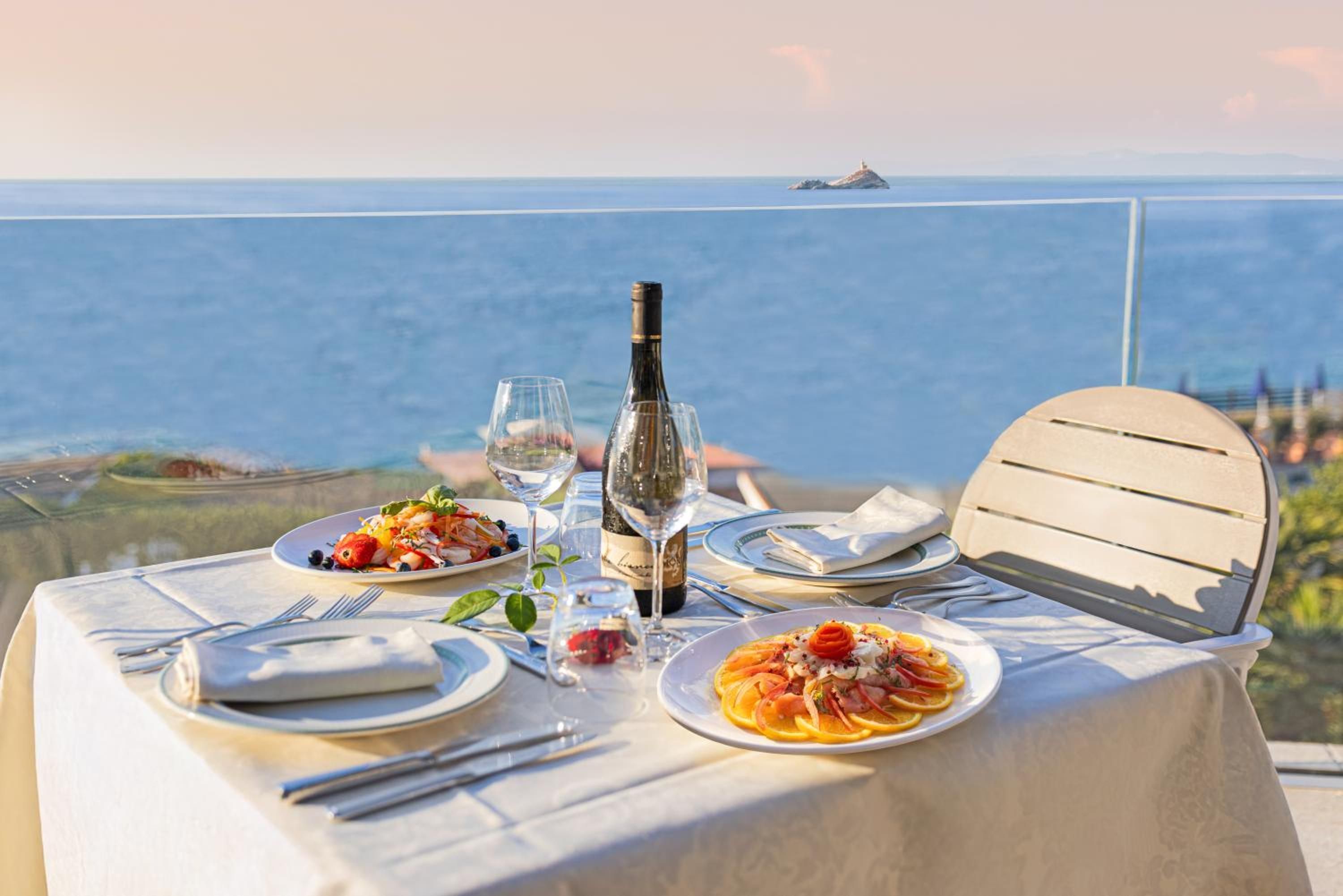 Un tavolo apparecchiato per due, con piatti di cibo, una bottiglia di vino e un bicchiere d'acqua, con vista su un'area lacustre e un'isola in lontananza.