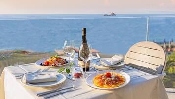 Un tavolo apparecchiato per due, con piatti di cibo, una bottiglia di vino e un bicchiere d'acqua, con vista su un'area lacustre e un'isola in lontananza.