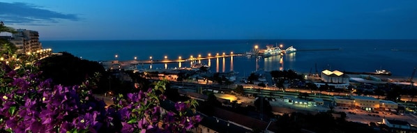 Un paisaje urbano costero al atardecer con edificios iluminados, un puerto deportivo y un puente.