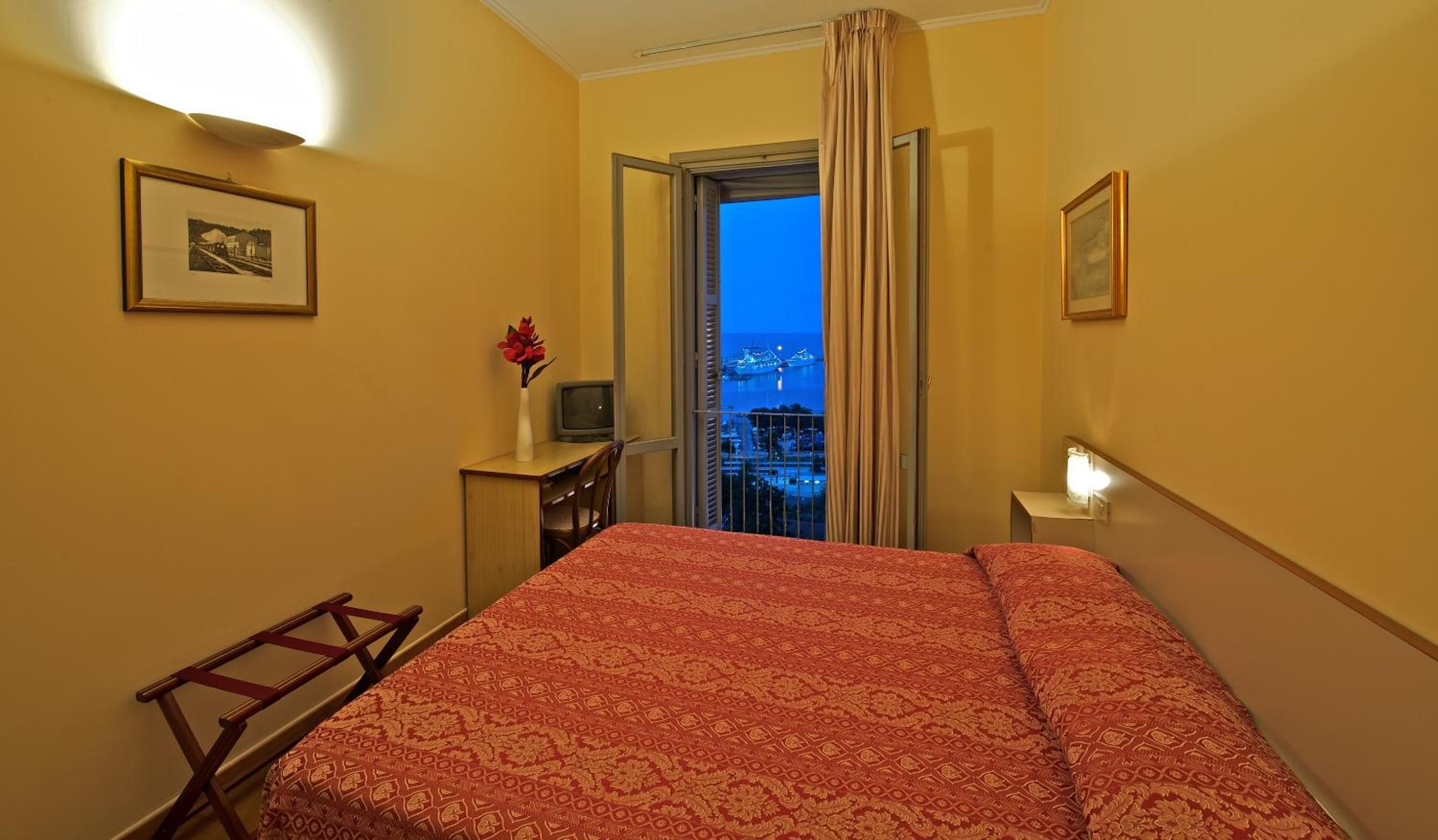 Una camera d'albergo con un letto, una scrivania, una sedia, una TV e una finestra con vista sulla città al crepuscolo.