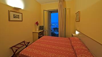 Una camera d'albergo con un letto, una scrivania, una sedia, una TV e una finestra con vista sulla città al crepuscolo.