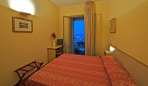 A hotel room with a bed, a desk, a chair, a TV, and a window with a view of a cityscape at dusk.