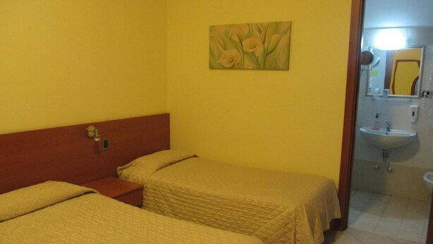 Habitación de hotel con dos camas, una mesita de noche, un cuadro en la pared y un baño con lavabo y espejo.