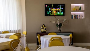 Un comedor con sillas y mesas amarillas, un televisor montado en la pared y objetos decorativos en estantes.