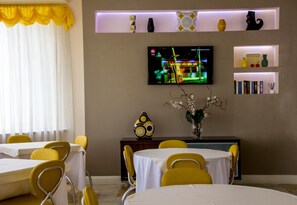 Un comedor con sillas y mesas amarillas, un televisor montado en la pared y objetos decorativos en estantes.