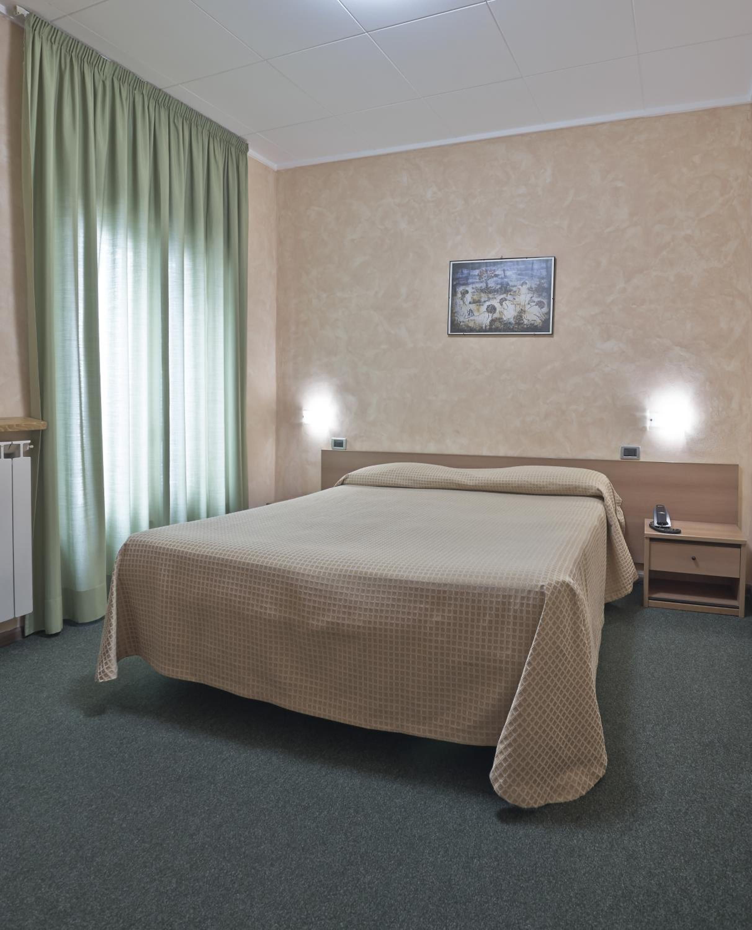 Camera d'albergo con un letto, un comodino, una lampada a parete, un termosifone e una finestra con tende.