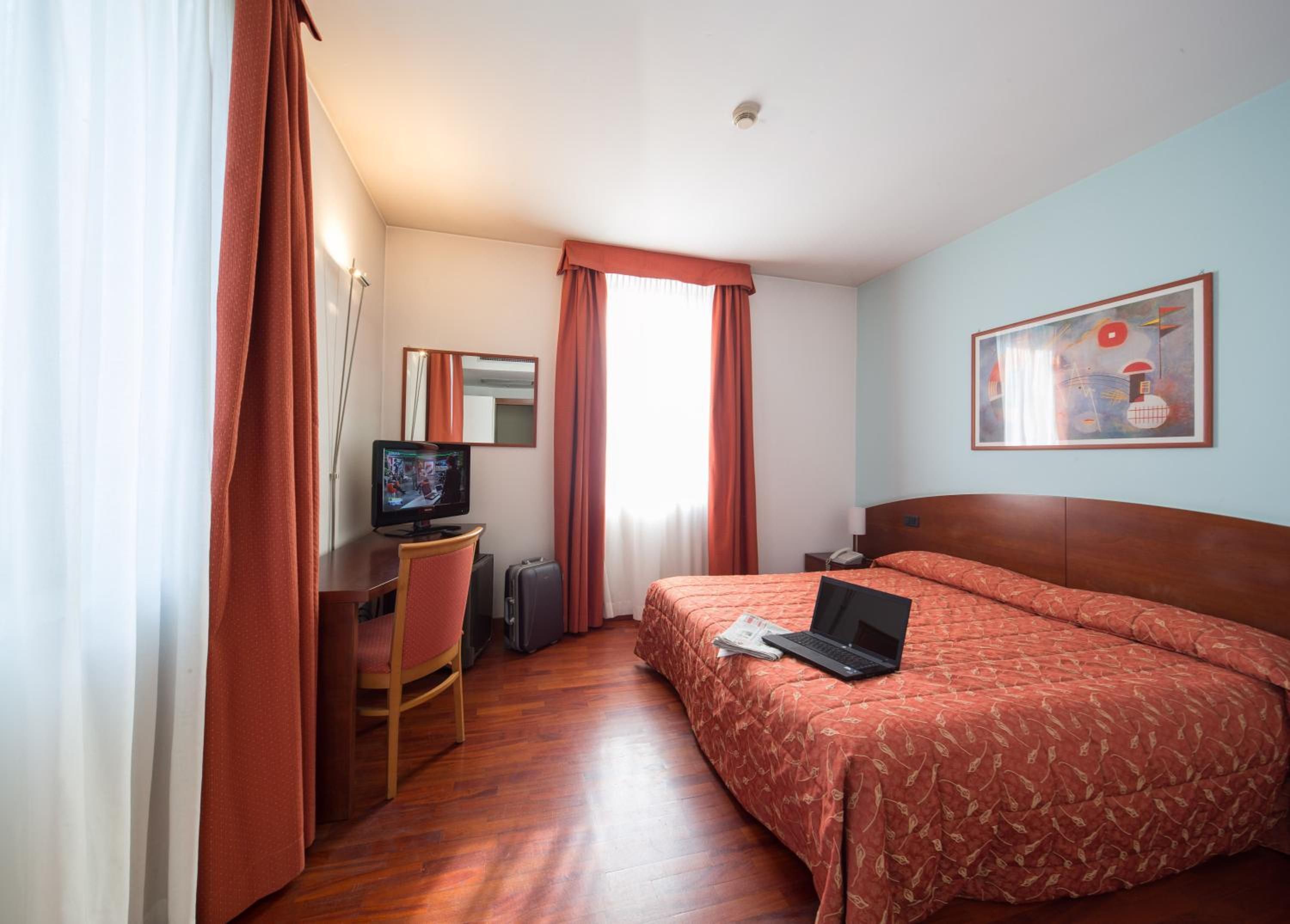 Una habitació d'hotel amb un llit, una taula de treball amb un ordinador, una cadira, una televisió i un quadre a la paret.