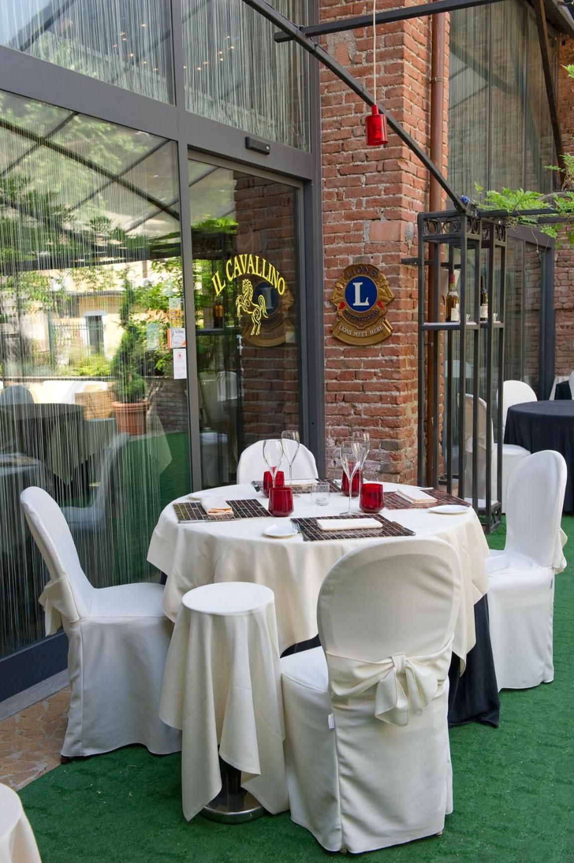 Un espacio exterior para comer con mesas redondas y sillas blancas, una pared de ladrillo y un ventanal con señalización.