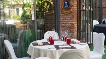 Un'area pranzo all'aperto con tavoli rotondi e sedie bianche, un muro in mattoni e una porta in vetro con insegne.