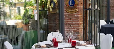 Un espacio exterior para comer con mesas redondas y sillas blancas, una pared de ladrillo y un ventanal con señalización.