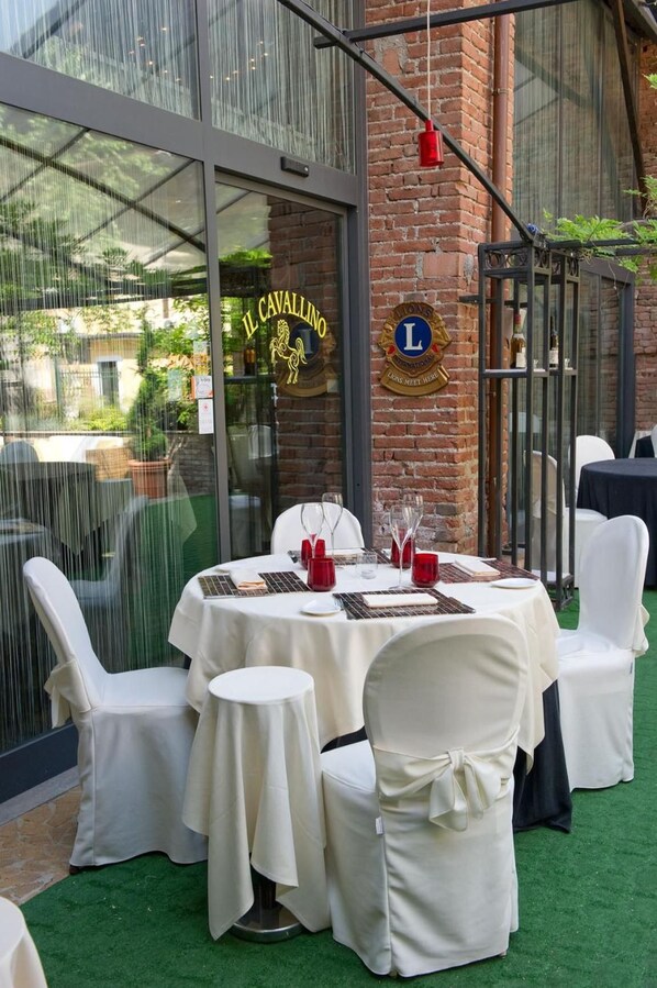 Un espacio exterior para comer con mesas redondas y sillas blancas, una pared de ladrillo y un ventanal con señalización.