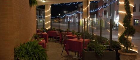 Un espace extérieur aménagé pour les repas, avec des tables et des chaises, des plantes en pot et des guirlandes lumineuses.