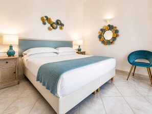 A neatly made bed with a blue throw, two bedside tables with lamps, a blue chair, and decorative wall art. - Hotel Guarracino (Capri)
