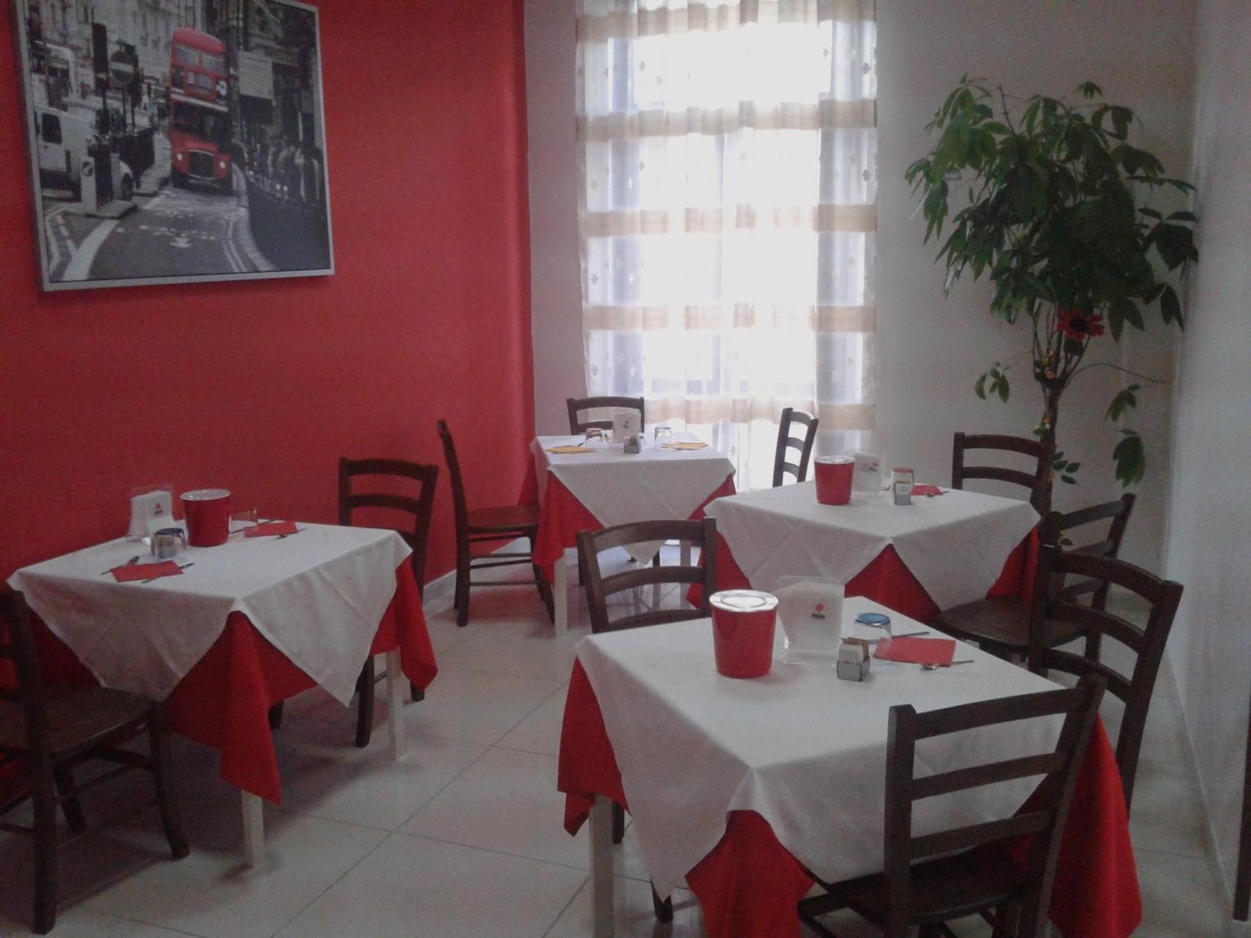Un comedor con mesas cubiertas con manteles blancos y servilletas rojas, sillas, una planta en maceta y un cuadro en la pared.